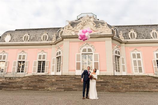 A groom and bride are Standing in front of a rose-painted Castle and are holding a few balloons.